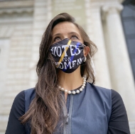 A female-appearing person with long, brown hair, pearl necklace, and blue top models the mask outside a white marble building.