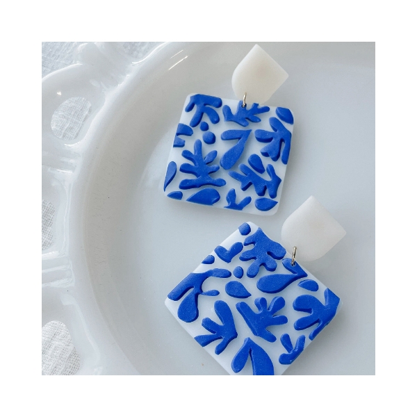 Two large, flat, square dangle earrings with blue abstract cutout designs fused to the white square surface. Handmade from polymer clay. 