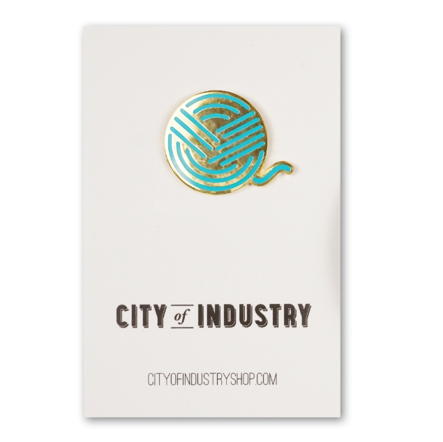 Round metal lapel pin mounted on a white card. It represents, in a stylized way, a ball of yarn. Gold chrome and blue-green enamel.