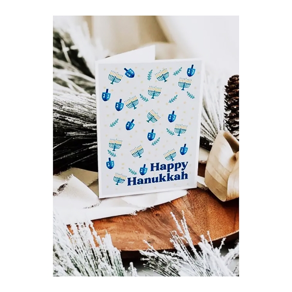 Happy Hanukkah Dreidel Card displayed on corner table surrounded by frosted pine needles and pine cones. Cover of card features white background with blue dreidels and menorahs scattered. 