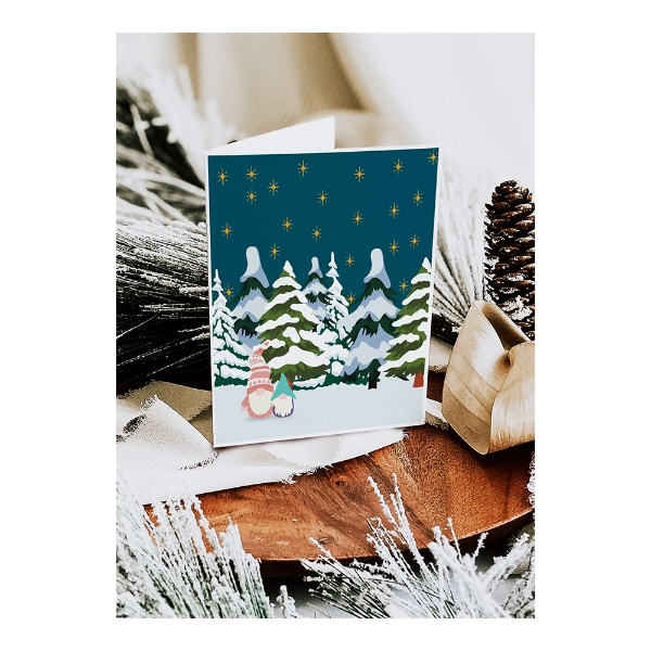 Gnome holiday card displayed on a table with frosted pine boughs and pine cones next to it. Card cover features illustration of 2 white bearded gnomes standing on snow in front of snow covered pine trees with a dark and starry sky. 